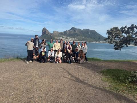 Large tour group poses at a coastal viewpoint overlooking dramatic mountains and a blue bay.