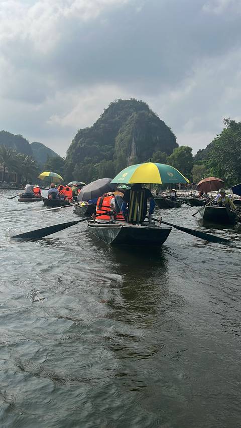 Tourists in orange life-jackets sit in metal rowboats with colourful umbrellas as they glide along a calm river flanked by lush limestone cliffs.