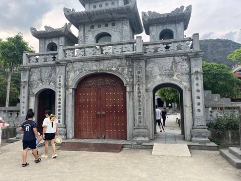 An ornate grey-stone pagoda gate with carved reliefs and wooden doors stands in a temple courtyard while visitors stroll past.