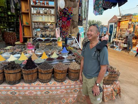 Smiling tourist stands beside colorful barrels and pyramid piles of spices at an Egyptian street market stall.