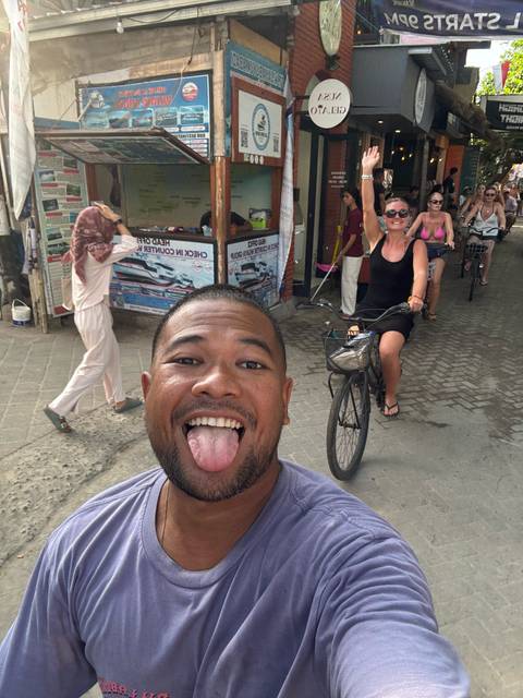 Selfie of excited cyclists and pedestrians along a narrow island street; lead traveler sticks out his tongue playfully.