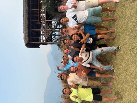 Travelers and local guides strike joyful poses on a grassy hillside with wooden stilt house and hazy volcanoes behind.