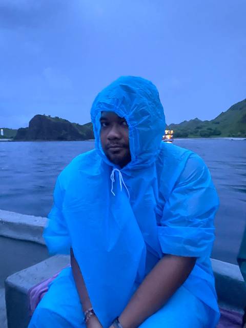 Man in blue rain poncho sitting on a boat at dusk with hilly shoreline behind.