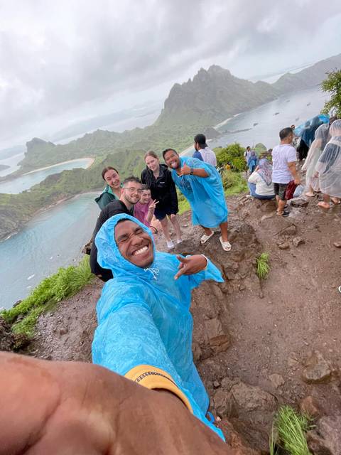 Wet but smiling hikers in blue ponchos posing on a coastal viewpoint overlooking bays and islands.