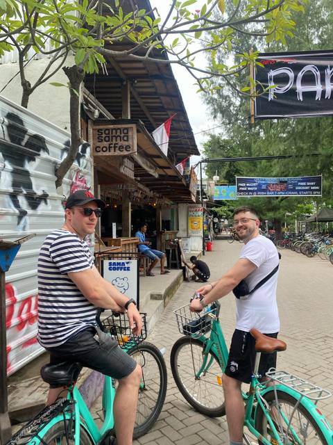 Two travelers with bicycles on a tropical island street lined with cafes and rental shops.
