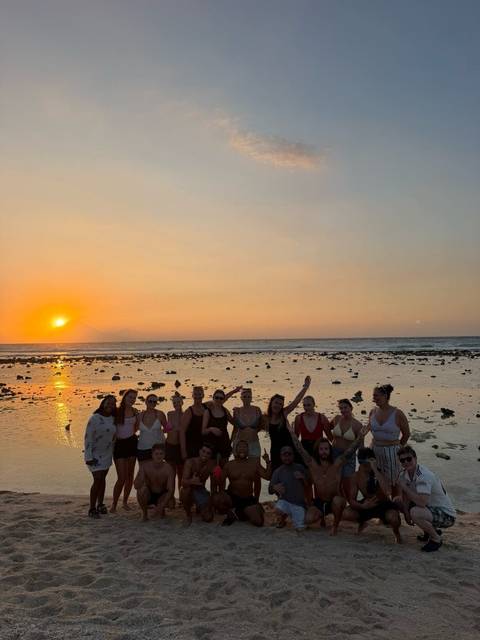 A line of young travelers posing on a rocky beach at sunset with the orange sun dropping over the sea horizon.