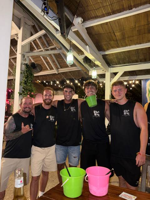 Five friends in matching black singlets smile at a lively bar decorated with string lights.