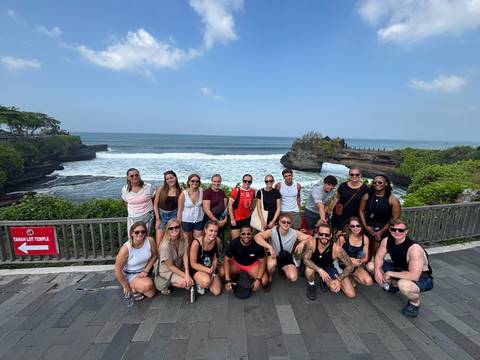 Tour group stands on a coastal boardwalk facing Tanah Lot Temple and crashing waves.
