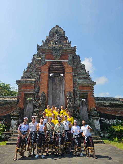 Participants in matching yellow shirts gather before an ornate Balinese split gate under blue sky.