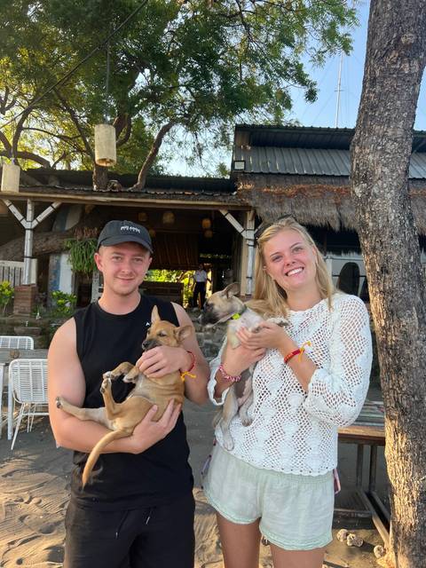 Smiling couple seated outdoors cuddle two small rescue puppies in front of a rustic café.