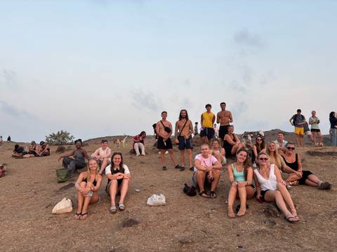Travelers sit on a dry hillside waiting for sunset with hazy sky overhead.