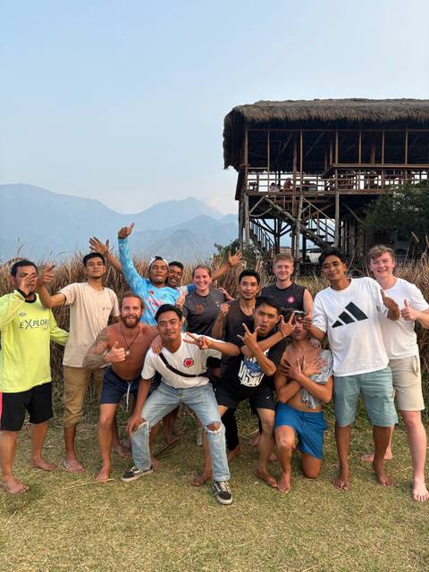 Tourists and local guides gesture playfully together beside a stilted wooden building near fields.