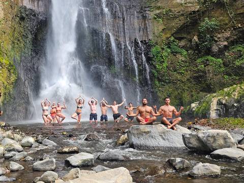 Adventurers strike yoga poses beneath a tall jungle waterfall, some standing in the pool, others meditating on rocks.