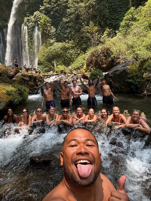 Group cools off in a rocky stream below a waterfall, leaning on the ledge and cheering to the camera.