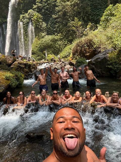Similar scene of friends enjoying a natural pool beneath a rainforest waterfall, posing for photos.
