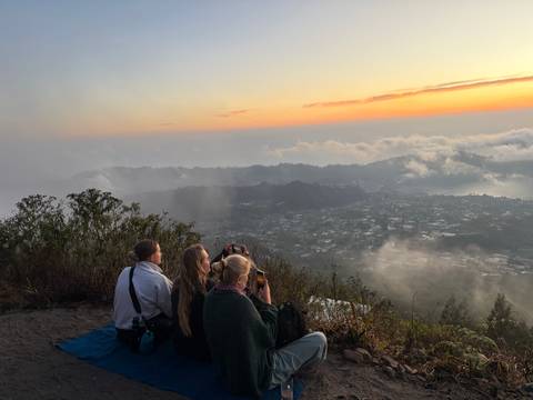 Three hikers sit on a ridge photographing a golden sunrise above misty valleys and a distant town.