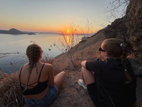 Two women watch a vivid sunset over calm island-dotted seas from a dry grassy slope.
