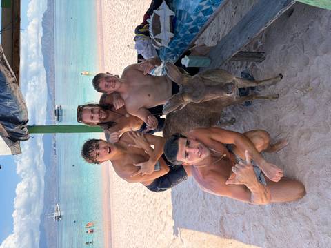 Four shirtless friends pose with a tame deer on a white-sand beach backed by turquoise water and boats.