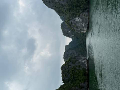 Narrow channel of emerald water flanked by towering forested karst cliffs beneath a cloudy sky.
