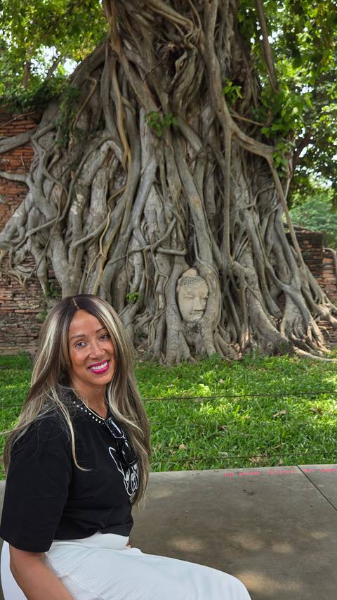 A woman sitting by a Buddha head embedded in a tree.