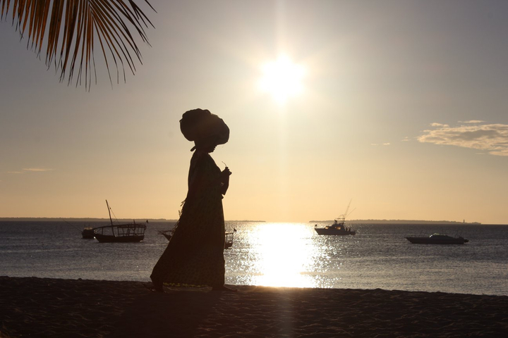 Silhouette of a person walking on the beach with boats on the water.