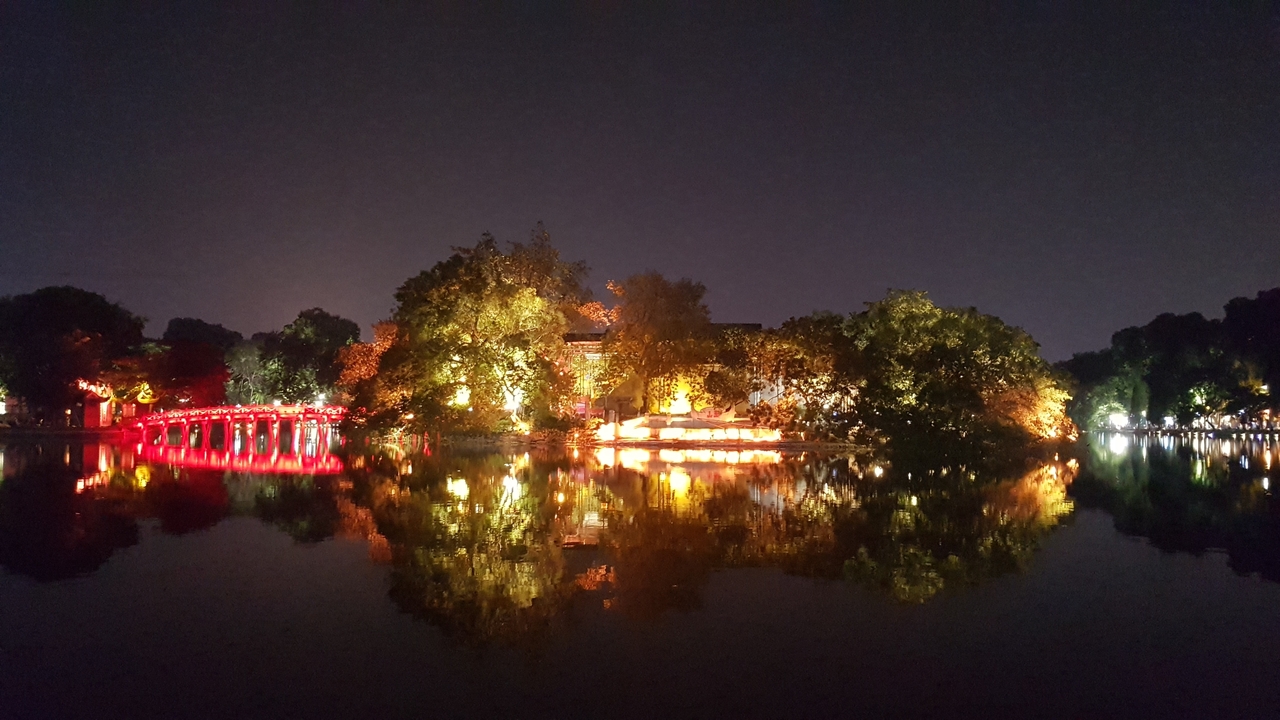 Pont et arbres illuminés de lumières vives se reflétant dans l'eau la nuit.