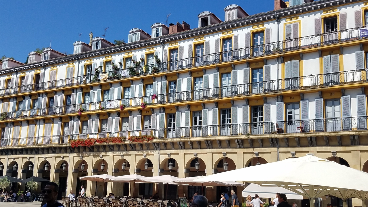 Rangée de bâtiments colorés avec des balcons de style français et une place ensoleillée.