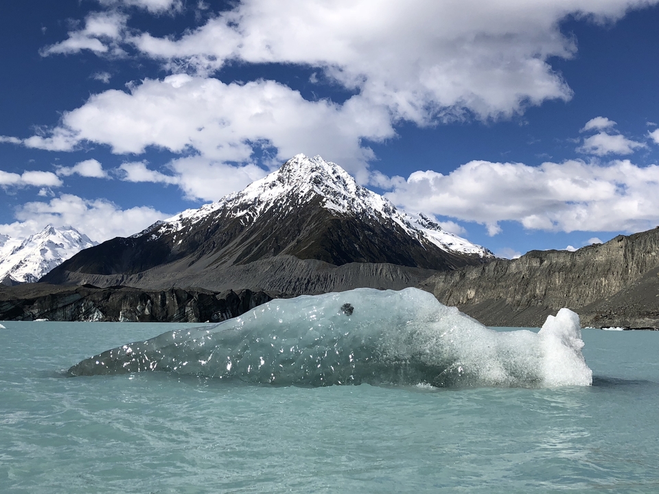 Iceberg in a turquoise lake with snow-capped mountains under blue sky.