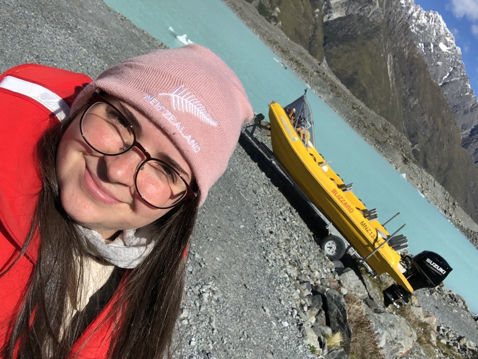 Une personne en veste rouge et chapeau rose souriante près d'un bateau jaune sur une plage rocailleuse.