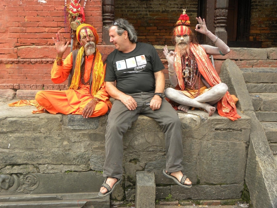Un homme assis entre deux sadhous vêtus traditionnellement avec un mur de briques en arrière-plan.