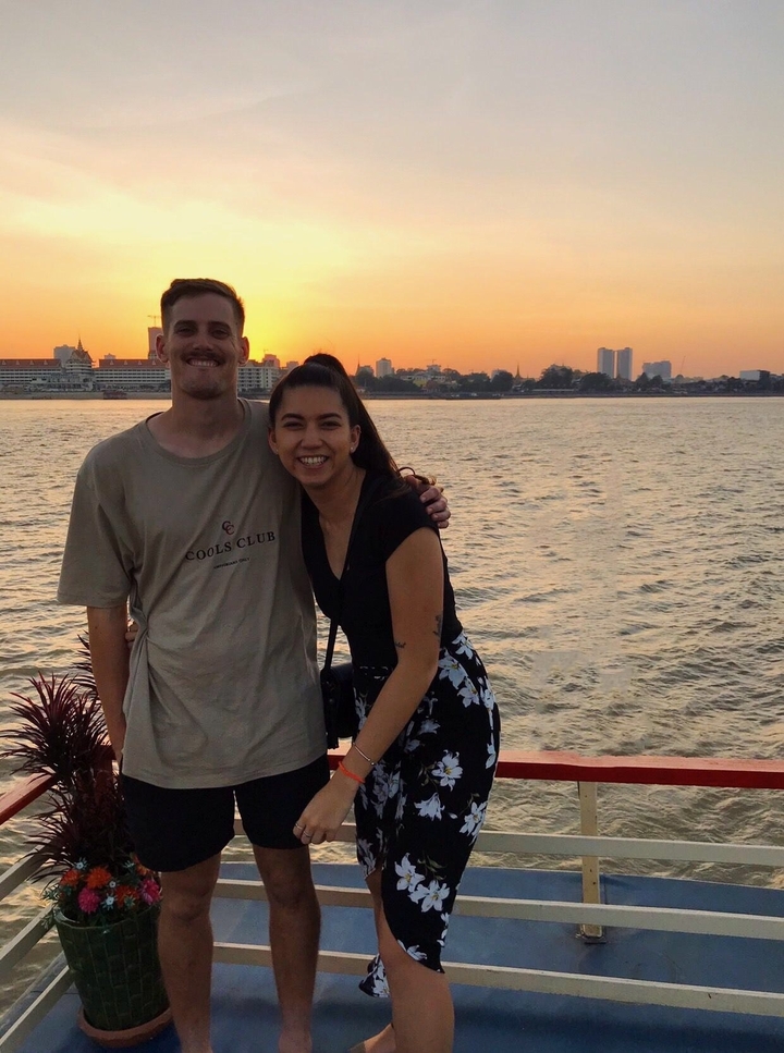 Couple posant près de l'eau avec un paysage urbain en arrière-plan au coucher du soleil.