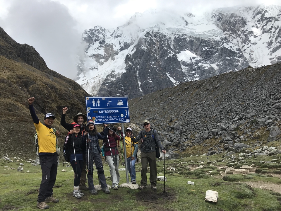 Groupe de randonneurs posant avec un panneau devant de majestueuses montagnes.