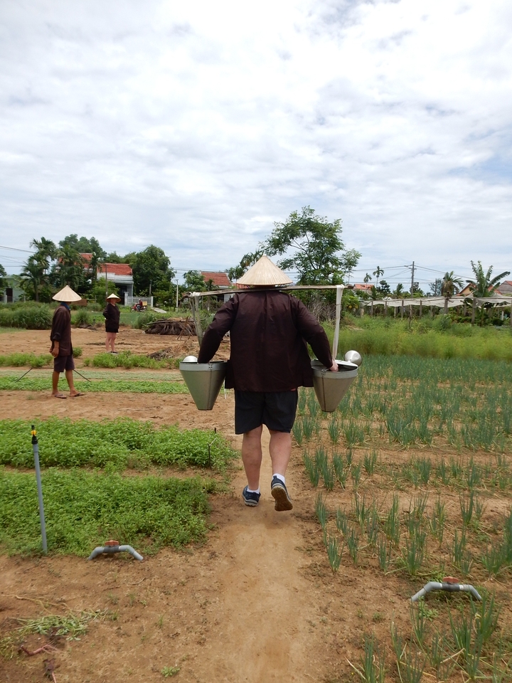 Personne portant un joug avec des arrosoirs dans un cadre rural.