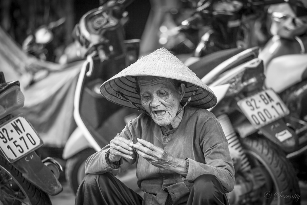Image en noir et blanc d'une personne âgée avec un chapeau traditionnel assise dans la rue.