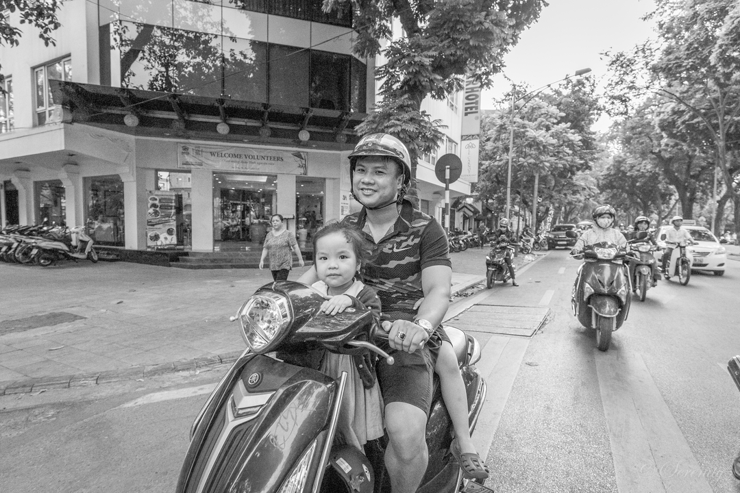 Image en noir et blanc d'un homme et d'un enfant sur un scooter dans une rue animée.