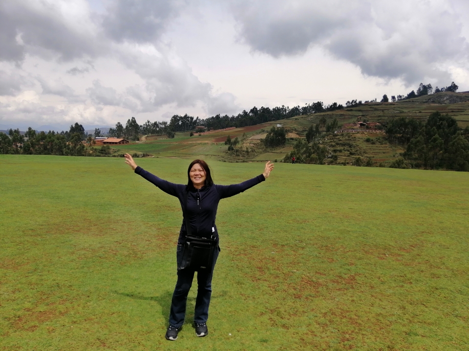 Une femme debout dans un champ herbeux, les bras étendus, avec des collines en arrière-plan.
