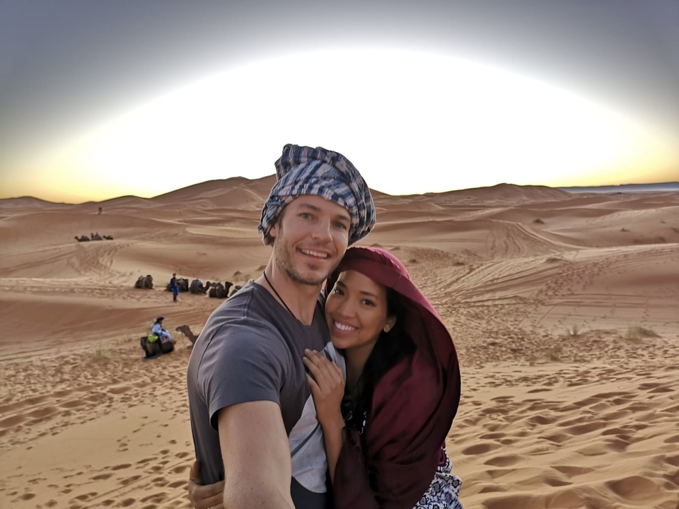 Couple souriant et posant sur des dunes du désert avec des chameaux en arrière-plan.