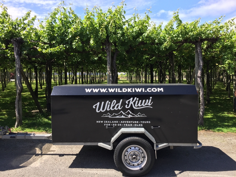 Gros plan d'une remorque avec le logo Wild Kiwi Tours garée dans un vignoble.