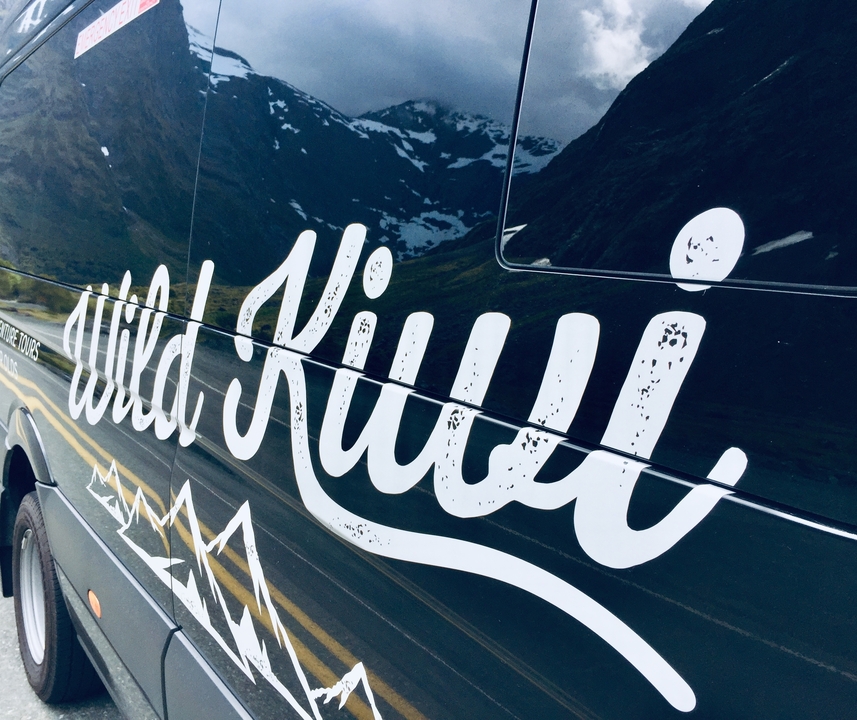 Fourgonnette avec « Wild Kiwi » peint dessus, garée sur une route à travers les montagnes.