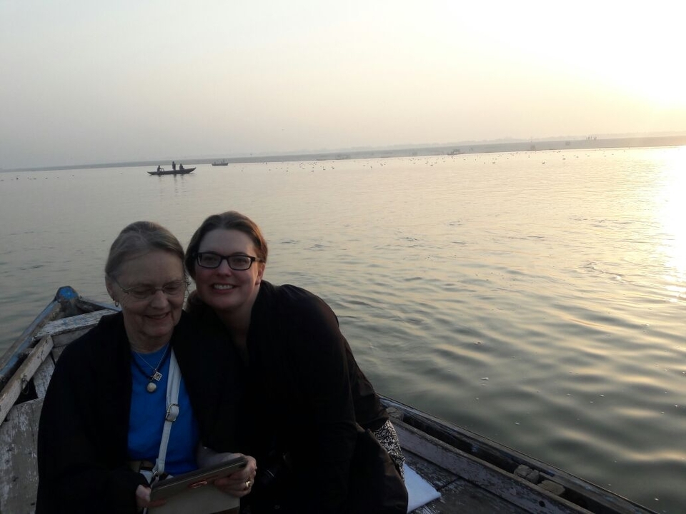 Two women sit on a wooden rowboat at dawn on a tranquil river with hazy sunlit horizon.