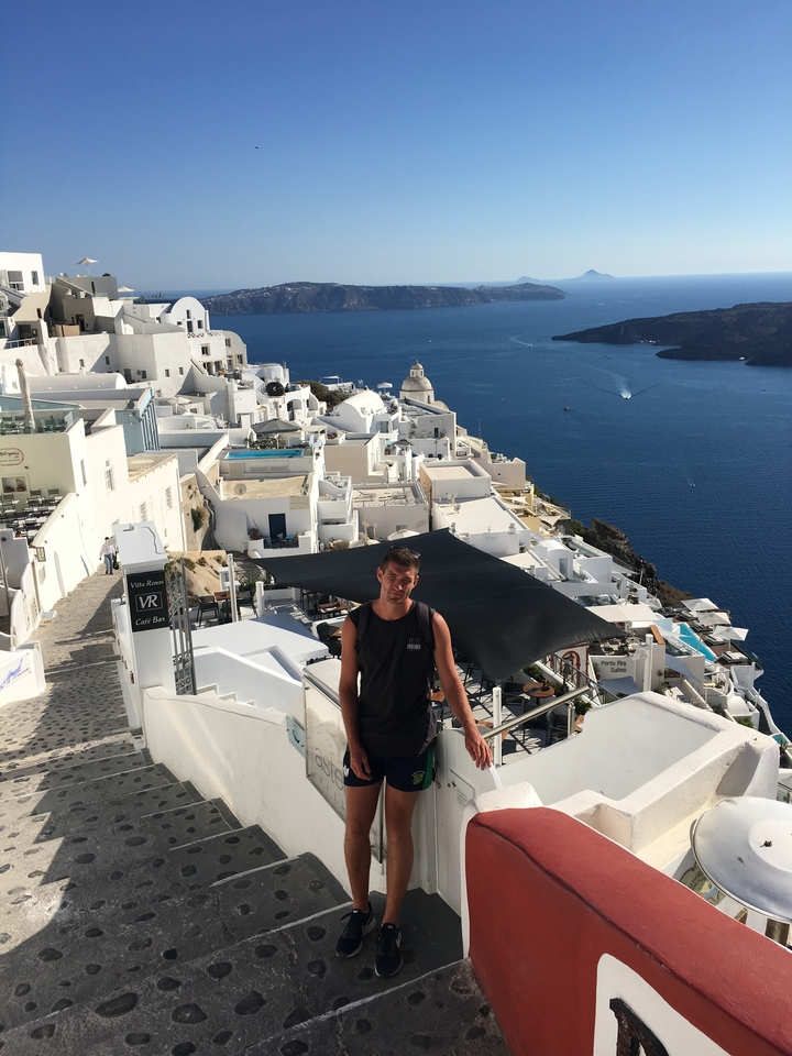 Person posing with a panoramic view of white-washed buildings and the sea.