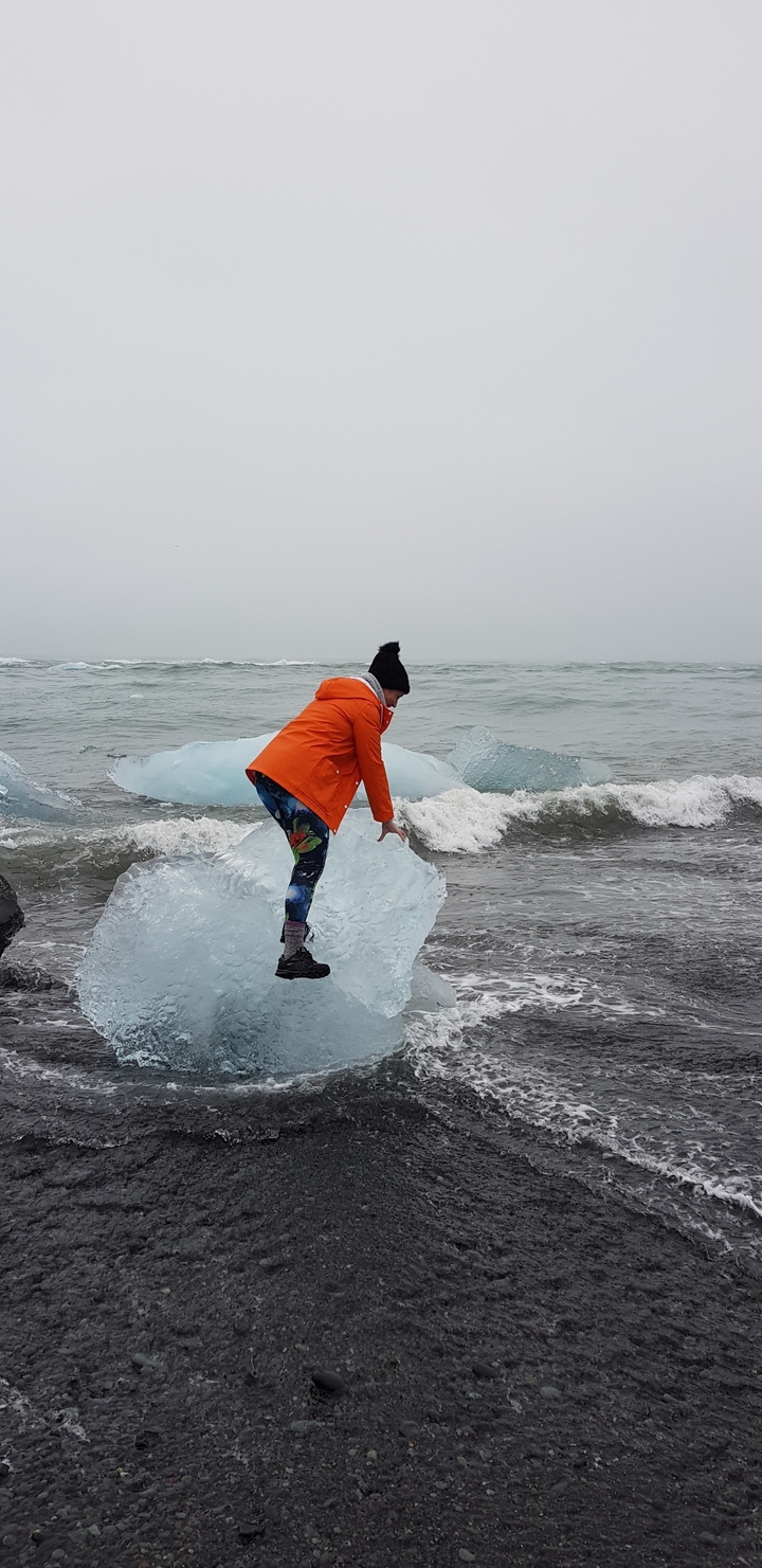 Personne en veste orange debout sur une plage interagissant avec des icebergs au bord de la mer.