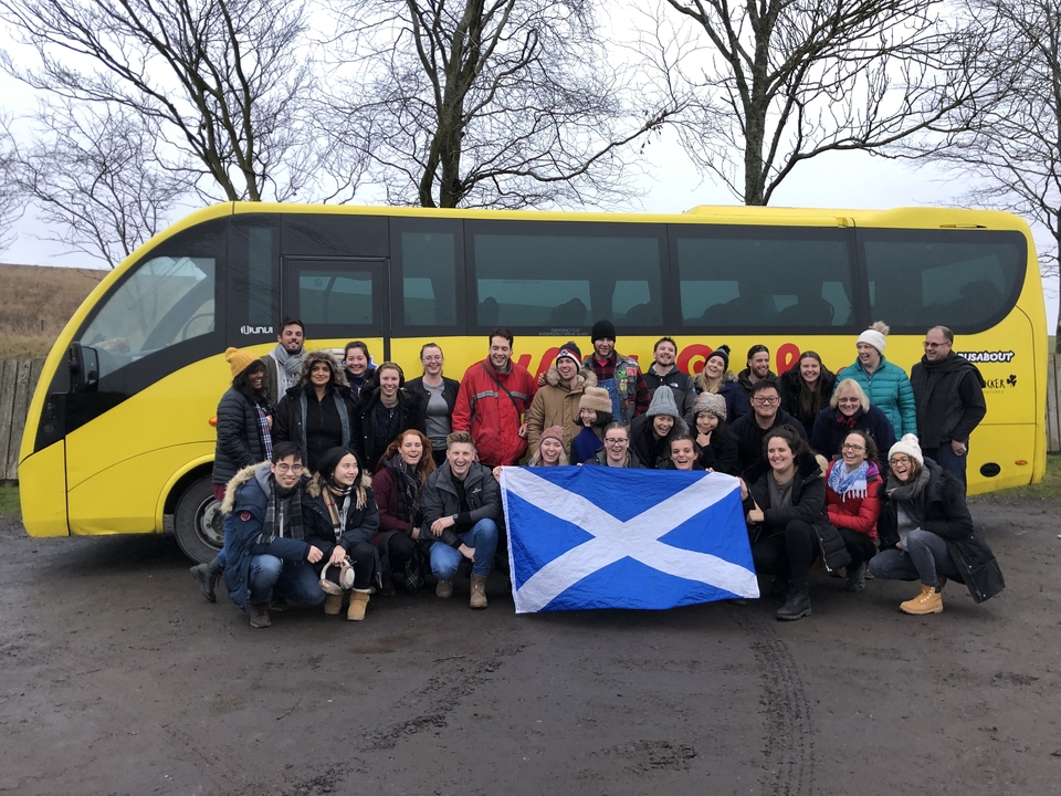 Grand groupe posant avec un drapeau écossais devant un bus jaune.