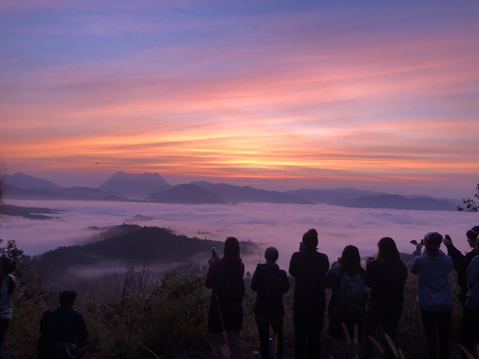 Des gens en silhouette contre un lever de soleil coloré au-dessus de montagnes brumeuses.