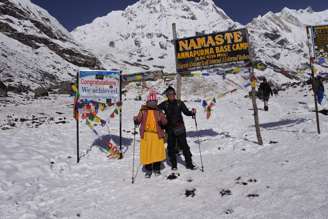 Deux personnes debout près de panneaux au camp de base de l'Annapurna avec des montagnes enneigées en arrière-plan.