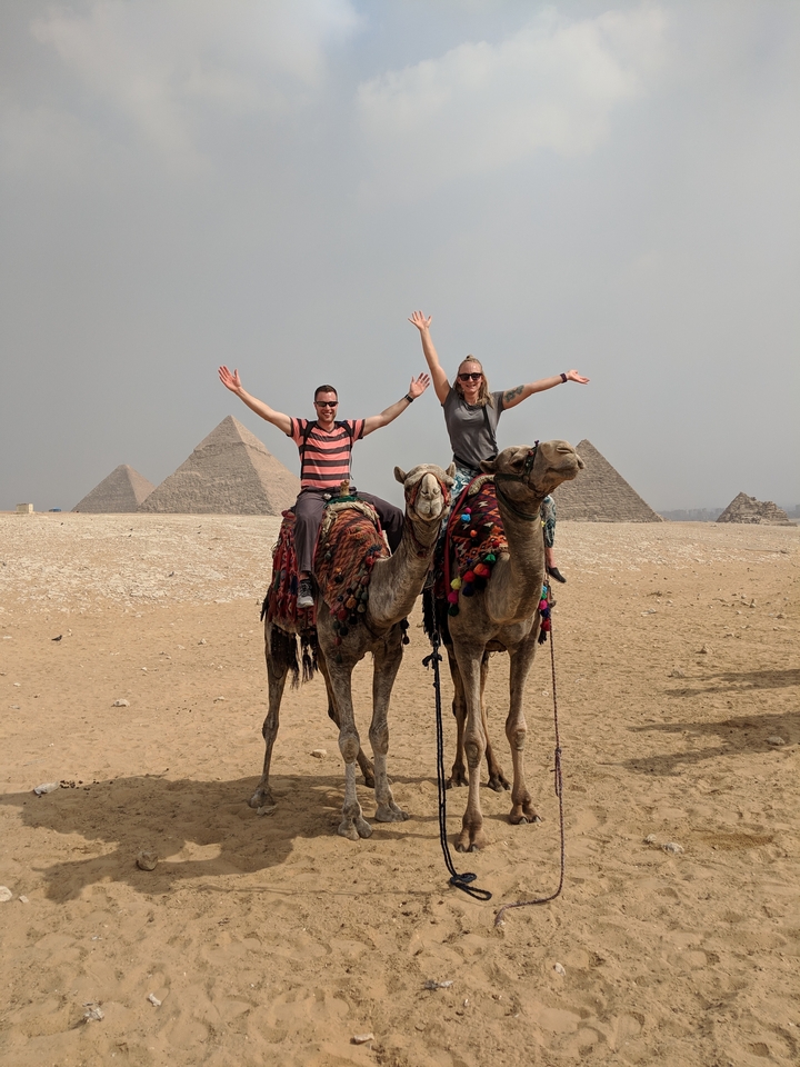Deux personnes à dos de chameau avec des pyramides en arrière-plan.