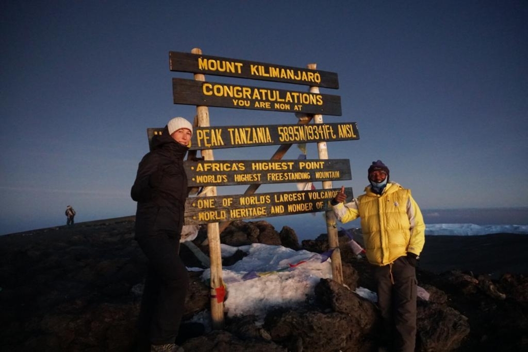 Deux personnes posant près du panneau du sommet sur le mont Kilimandjaro à l'aube.
