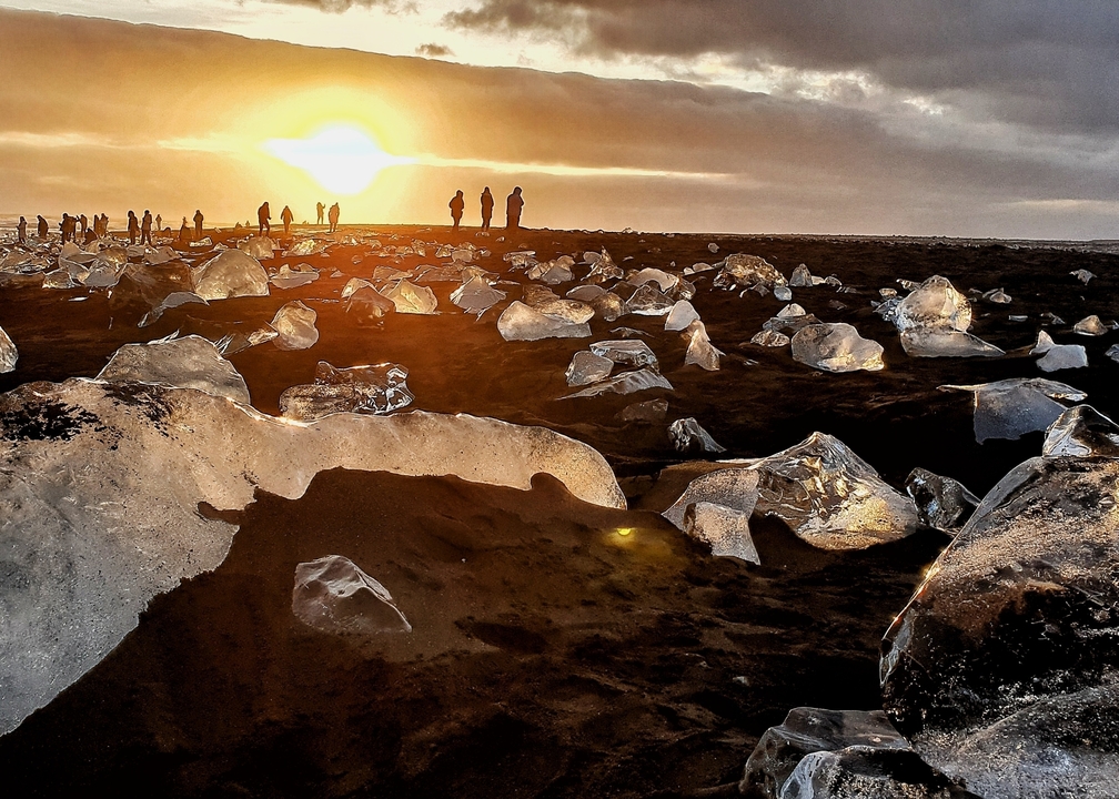 Vue du coucher de soleil sur une plage de sable noir avec des blocs de glace et des silhouettes de personnes.