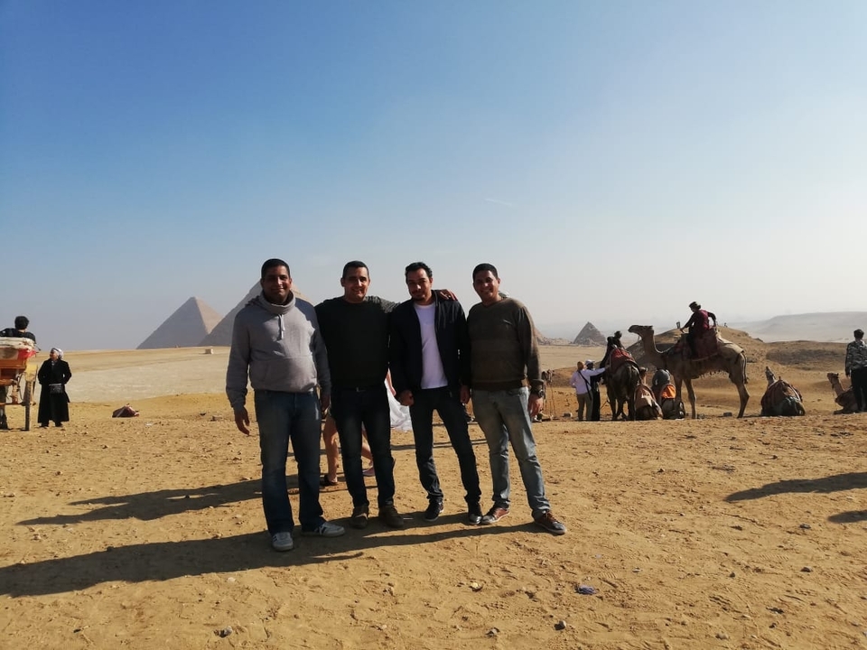 Groupe d'hommes posant avec des pyramides et un chameau en arrière-plan.