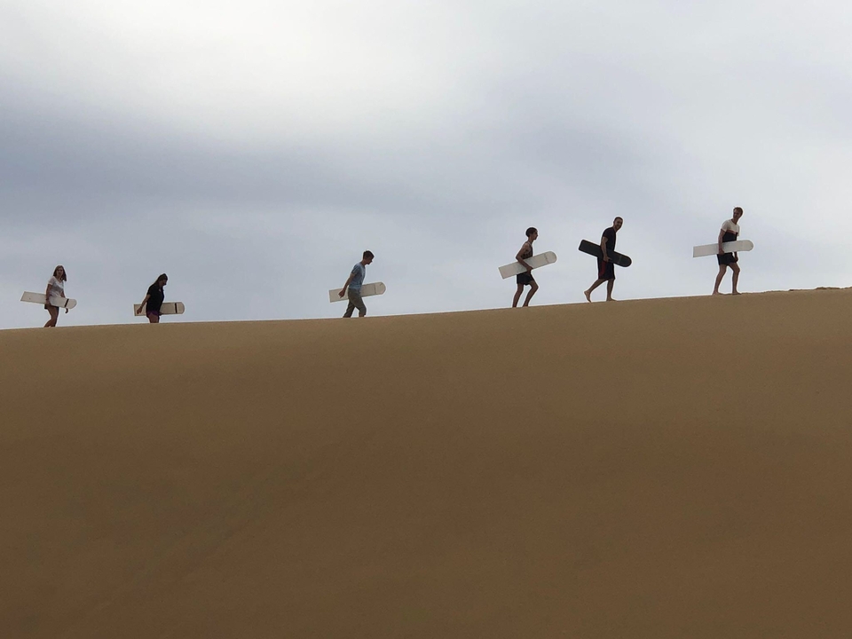 People carrying boards in sand dunes.